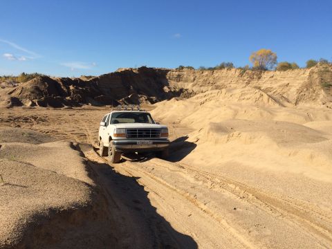 1993 Ford Bronco - A white Ford Bronco drives through a sandy quarry under a clear blue sky. The SUV has four auxiliary lights mounted on its roof.