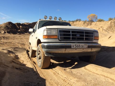 1993 Ford Bronco - A white 1993 Ford Bronco is parked on a sandy incline under a clear blue sky. The front of the SUV is facing the camera, with off-road ligh…