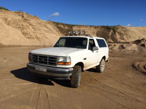 1993 Ford Bronco - A white 1990s Ford Bronco is parked on a sandy terrain with large sand dunes in the background under a clear blue sky. The vehicle has four…