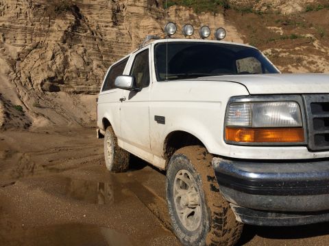 1993 Ford Bronco - A white 1993 Ford Bronco is parked on a muddy, wet surface with a sandy cliff face in the background. The vehicle is viewed from the front…