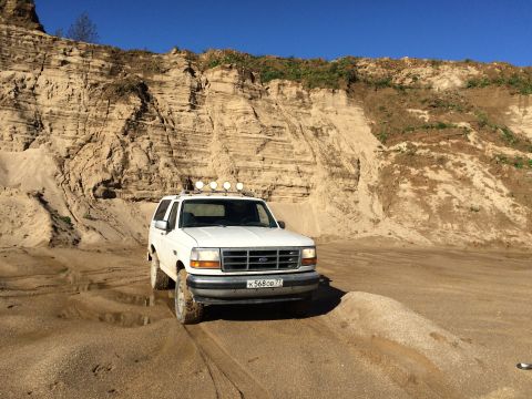 1993 Ford Bronco - A white Ford Bronco is parked on a sandy terrain with a large sand dune in the background under a clear blue sky. The vehicle has four auxi…
