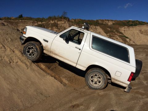 1993 Ford Bronco - A white 1993 Ford Bronco is shown at an angle, driving up a sandy incline with a dirt cliff face in the background under a clear blue sky.