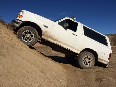 1993 Ford Bronco - A white Ford Bronco is driving up a steep sandy embankment under a clear blue sky. The vehicle is tilted at an angle, with mud splattered o…
