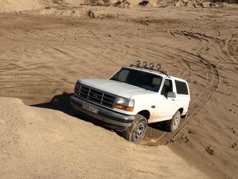 1993 Ford Bronco - A white Ford Bronco is driving on a sandy dune, with its front tires partially sunk into the sand. The vehicle has four auxiliary lights mo…