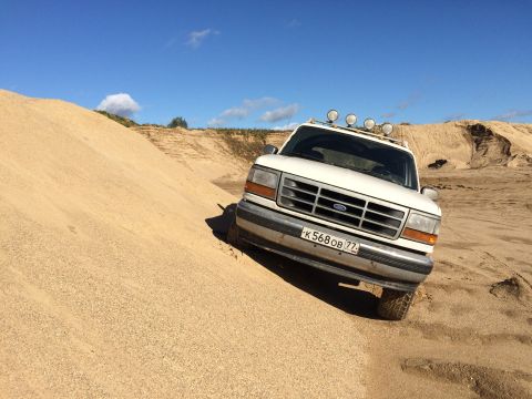1993 Ford Bronco - A white Ford Bronco is parked on a sandy incline in a desert-like environment under a clear blue sky. The vehicle has four auxiliary lights…