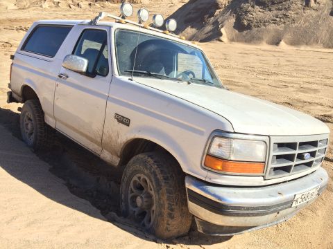 1993 Ford Bronco - A white 1993 Ford Bronco is stuck in the sand, with its front tires partially buried. The vehicle has a roof rack with four auxiliary light…