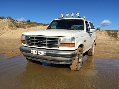 1993 Ford Bronco - A white 1993 Ford Bronco is parked in shallow water and mud with sand dunes in the background under a clear blue sky. The vehicle has four…