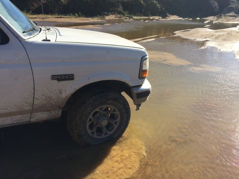 1993 Ford Bronco - The front quarter panel and tire of a white Ford Bronco are submerged in shallow, muddy water. The vehicle is off-road, with dirt and mud s…