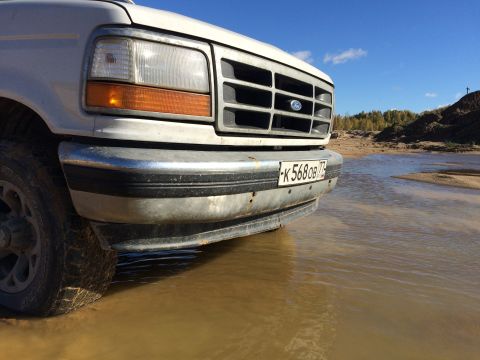 1993 Ford Bronco - A white Ford Bronco is partially submerged in shallow, muddy water, with its front grille and bumper clearly visible. The vehicle is parked…
