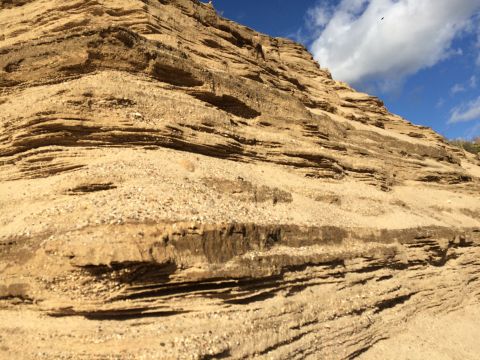 1993 Ford Bronco - A low-angle shot of a sandy cliff face with distinct horizontal layers of sediment and some gravel. The sky above is blue with scattered wh…