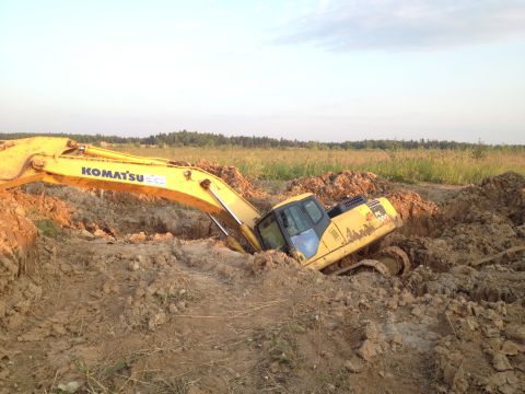1993 Ford Bronco - A yellow Komatsu excavator is stuck in a deep dirt trench, with its boom arm extended and the cab tilted. The surrounding area is a constru…