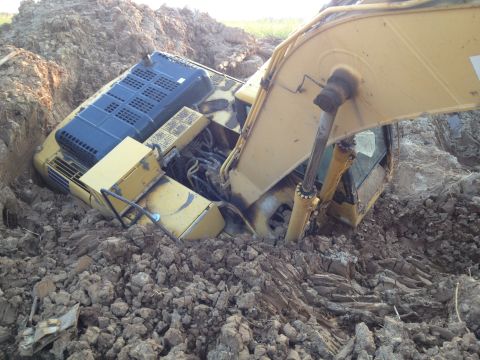 1993 Ford Bronco - A yellow excavator is stuck in a muddy trench. The machine is partially buried in dirt and appears to be at an angle.