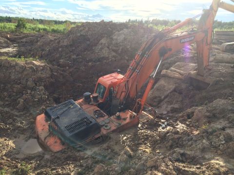 1993 Ford Bronco - An orange excavator is stuck in a muddy pit, with its tracks submerged in water and mud. The excavator is tilted to the side, with its boom…