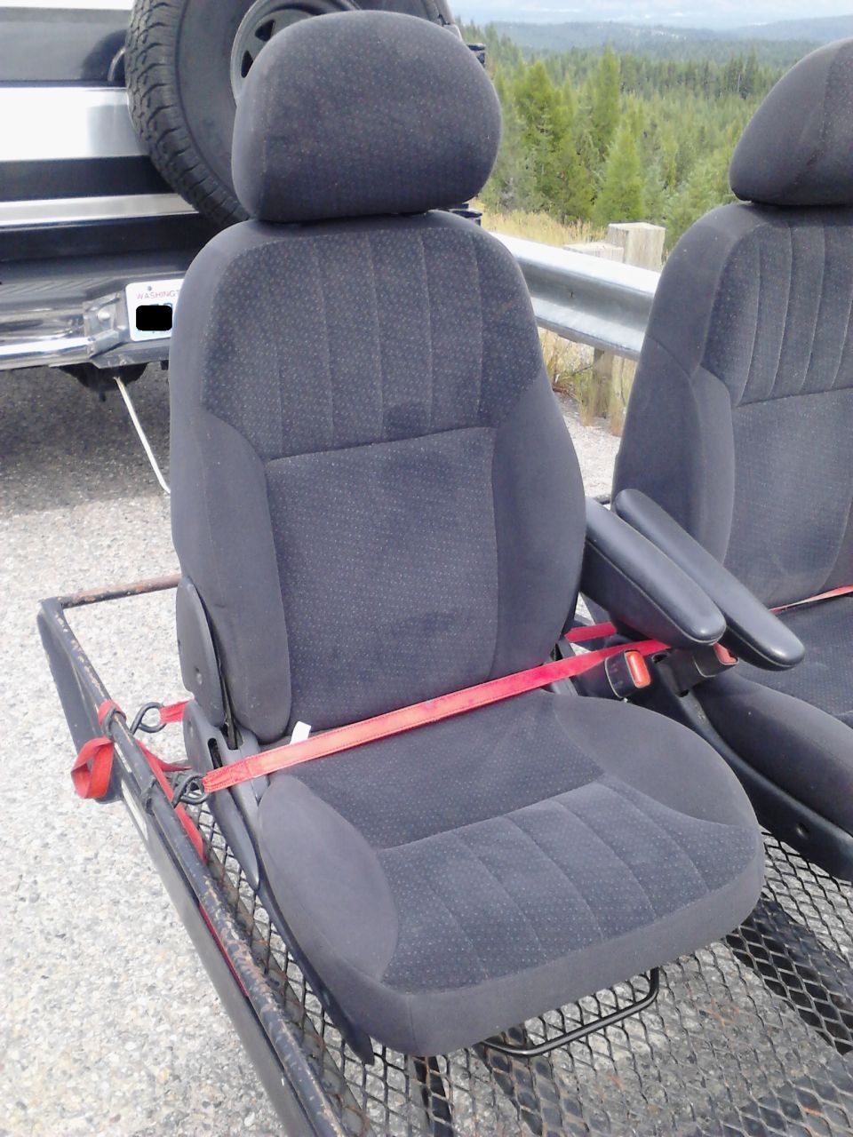 Two dark gray car seats strapped to a trailer, with a tire and vehicle in the background.