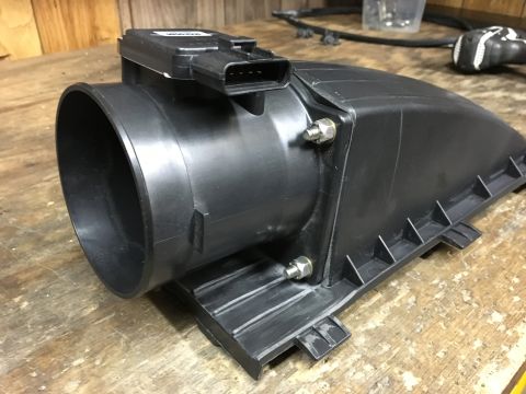 1996 Ford Bronco - A black plastic mass air flow sensor housing with a sensor attached is shown on a wooden surface. The housing has a large circular intake o…