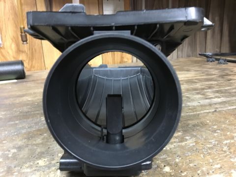 1996 Ford Bronco - A black plastic mass airflow sensor housing is shown on a wooden workbench. The housing has a large circular opening with internal fins and…