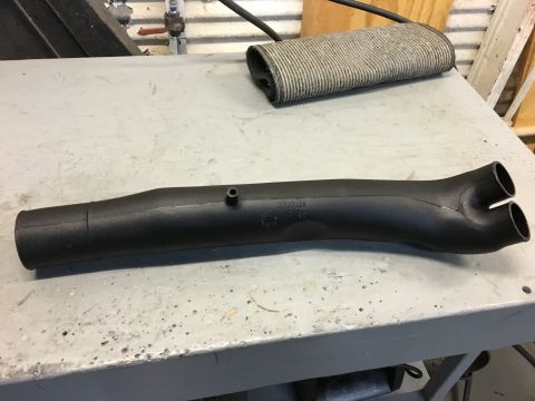 1996 Ford Bronco - A black, cast metal intake manifold with a Y-shaped end is shown resting on a grey workbench. The manifold has a small raised nipple and so…