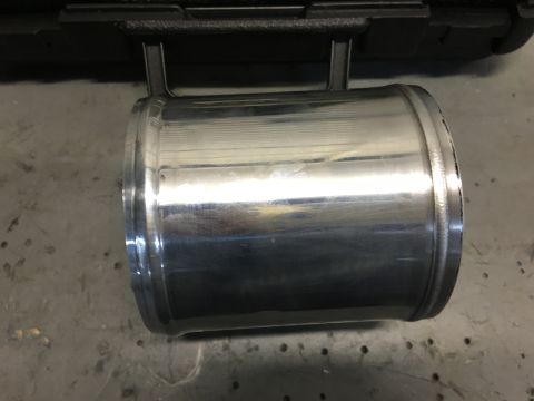 1996 Ford Bronco - A polished aluminum intake pipe with rolled ends sits on a textured gray surface, with a black object in the background.