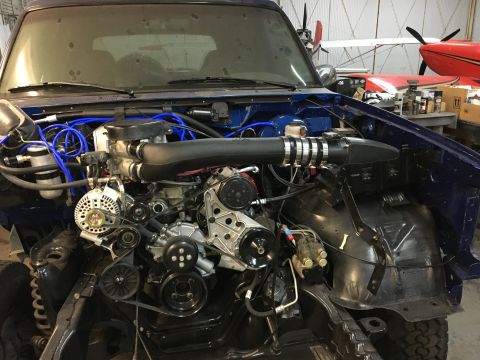 1996 Ford Bronco - The engine bay of a dark blue 1996 Ford Bronco is shown with the hood removed, revealing the engine, alternator, and various hoses and wiri…