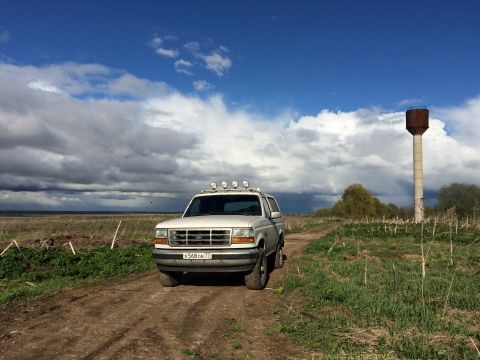 1993 Ford Bronco - A white 1993 Ford Bronco is parked on a dirt road in a grassy field under a cloudy blue sky. A tall water tower stands in the background.