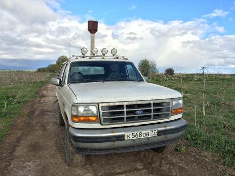 1993 Ford Bronco - A white 1993 Ford Bronco is parked on a dirt road in a grassy field under a cloudy sky. The vehicle has a roof rack with four auxiliary lig…