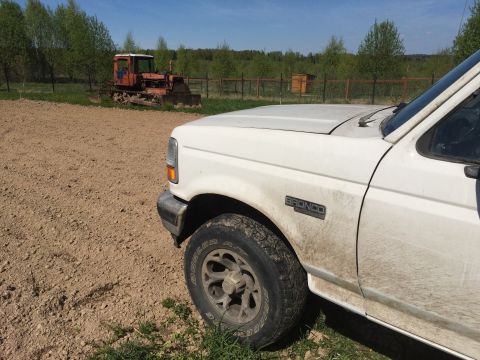 1993 Ford Bronco - The front quarter panel and tire of a white Ford Bronco are visible, covered in dirt. In the background, an orange tractor sits in a field…