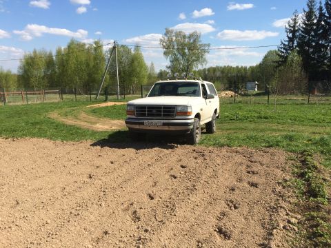 1993 Ford Bronco - A white 1993 Ford Bronco is parked on a dirt patch in a grassy field with trees in the background. The vehicle has four off-road lights mou…
