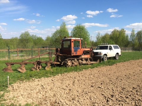 1993 Ford Bronco - A white 1990s Ford Bronco SUV is parked next to a rusty orange tractor with a plow attachment in a grassy field with a plowed section in th…