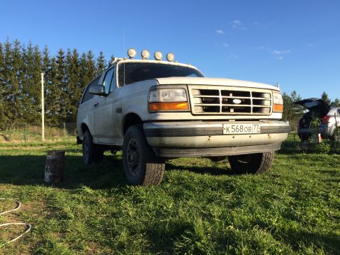 1993 Ford Bronco - A white 1993 Ford Bronco with four auxiliary lights mounted on the roof rack is parked on a grassy field with a line of pine trees in the b…