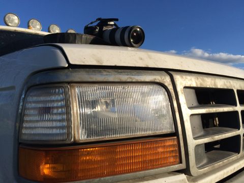1993 Ford Bronco - A close-up, low-angle shot of the front of a white Ford Bronco shows its headlights, grille, and hood. A Canon camera with a large lens res…