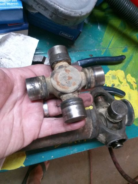 1996 Ford Bronco - A hand holds a U-joint assembly with visible rust and wear. Another U-joint is attached to a driveshaft in the background.