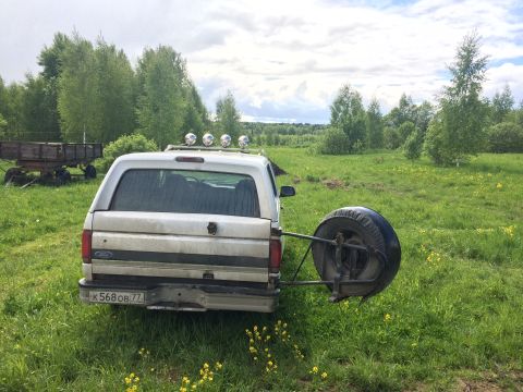 1993 Ford Bronco - The rear view of a white Ford Bronco is shown parked in a grassy field with a spare tire mounted on the back. A wooden trailer is visible t…