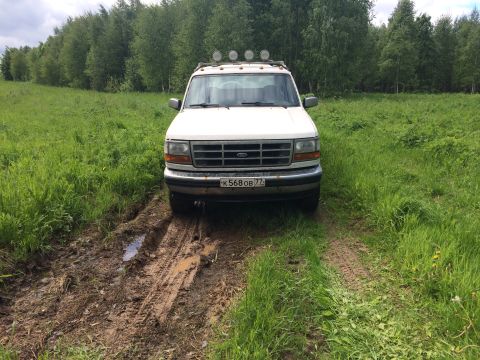 1993 Ford Bronco - A white Ford Bronco is parked on a muddy dirt road in a grassy field with trees in the background. The vehicle has a roof rack with lights…