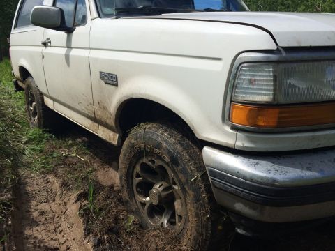 1993 Ford Bronco - A white Ford Bronco is stuck in mud on a grassy trail. The front passenger side tire and fender are visible, along with the headlight and b…