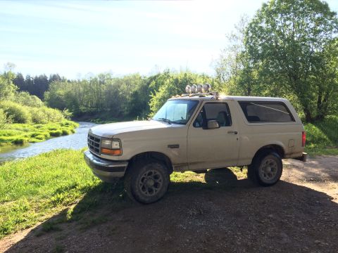 1993 Ford Bronco - A tan 1993 Ford Bronco is parked on a dirt path next to a winding river and lush green trees. The SUV has four auxiliary lights mounted on…