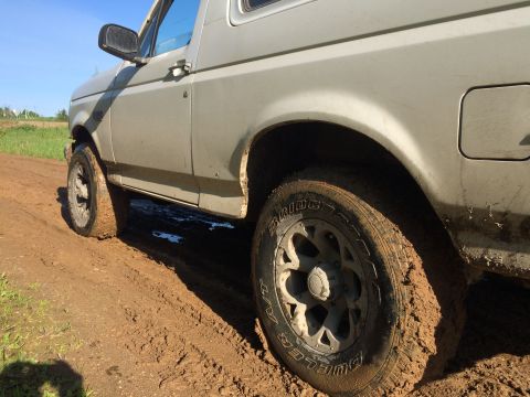 1993 Ford Bronco - A silver Ford Bronco is driving through a muddy field, with its tires covered in mud. The sky is clear and blue.