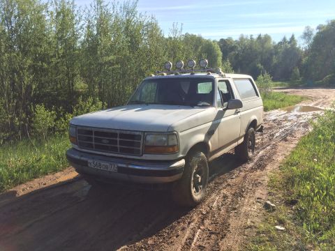 1993 Ford Bronco - A white 1993 Ford Bronco is parked on a muddy dirt road surrounded by trees. The vehicle has a roof rack with four auxiliary lights and is…