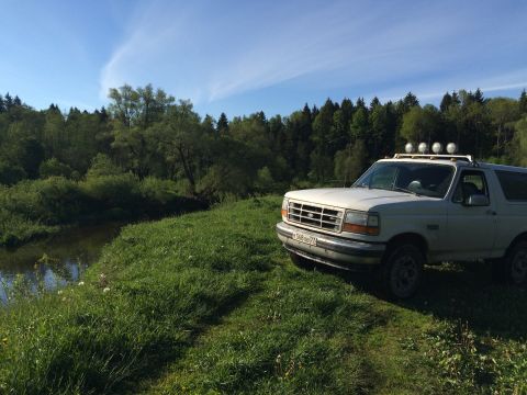 1993 Ford Bronco - A white 1993 Ford Bronco is parked on a grassy hill next to a river, with a dense forest in the background under a blue sky with wispy clou…
