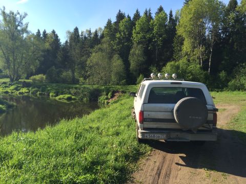 1993 Ford Bronco - A white Ford Bronco is parked on a dirt road next to a grassy riverbank with a dense forest in the background.