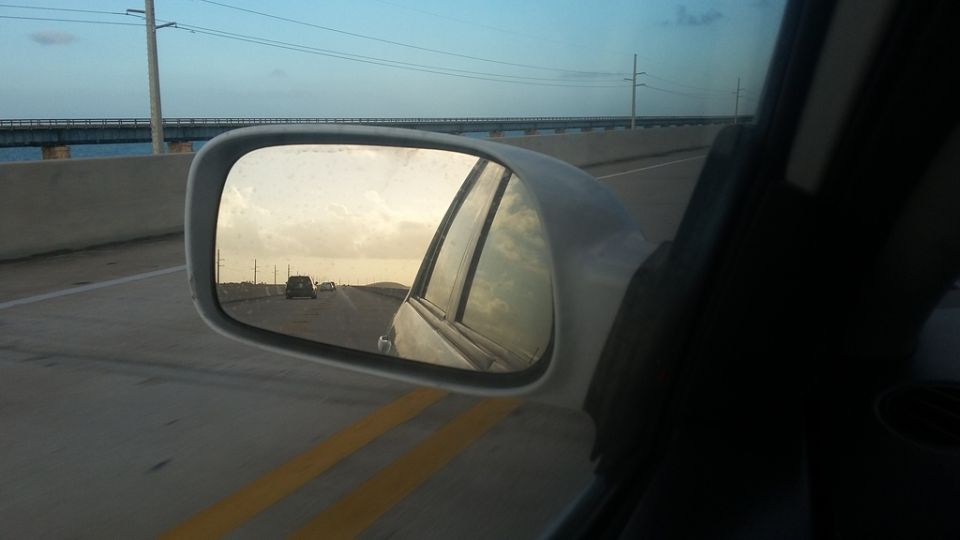 Car side mirror view of a highway overpass and cars on a road at sunset.