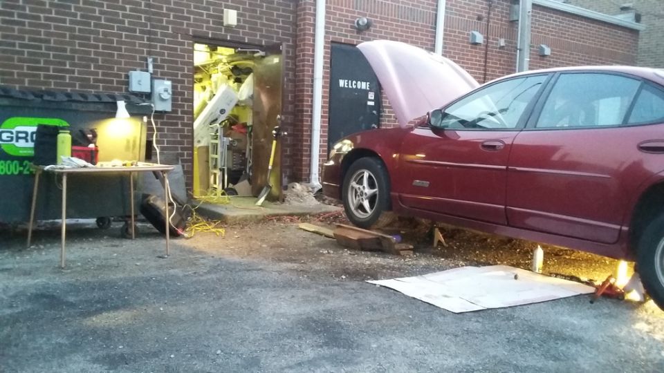 Maroon Pontiac Grand Prix sedan on jack stands with hood open, next to a dumpster and a table with tools.