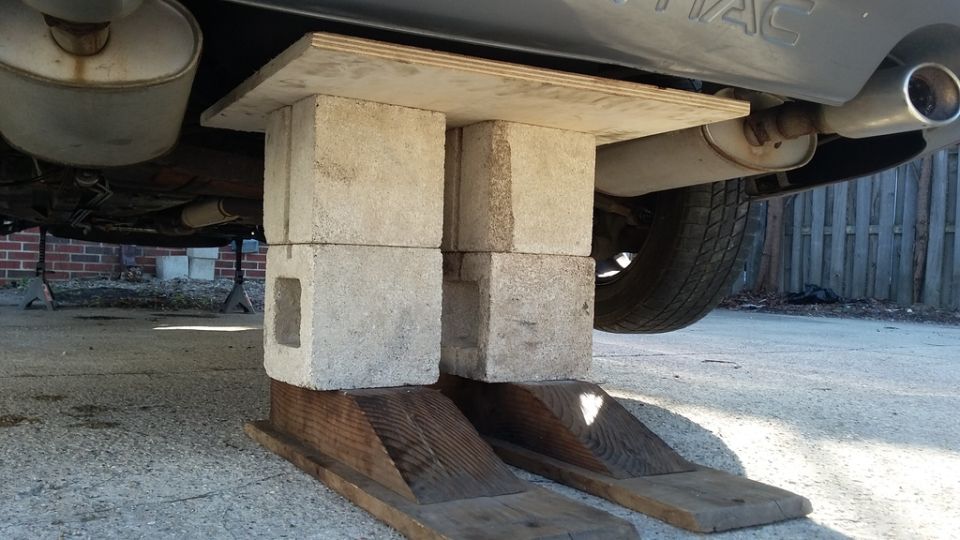 Underside of a car supported by cinder blocks and wooden ramps, showing exhaust and tire.