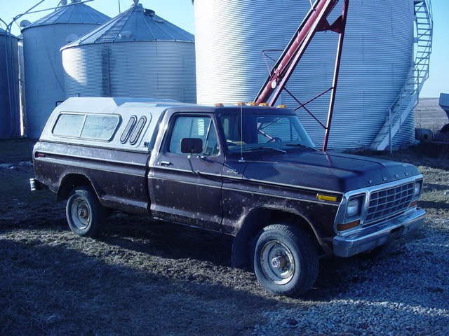 Brown Ford F-Series pickup truck with a white camper shell, parked in front of grain silos