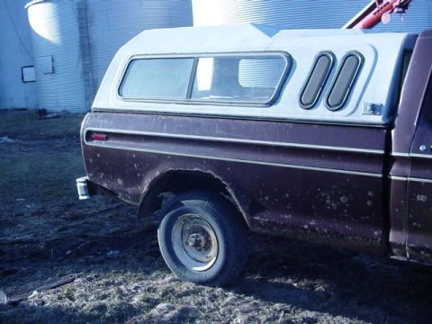 1992 Ford Bronco - A pickup truck with a camper shell is parked outdoors near large silos. The truck appears to be older and shows some wear.