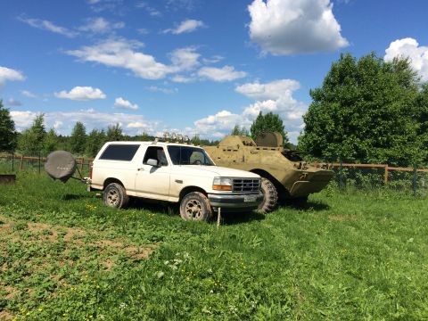1993 Ford Bronco - A white Ford Bronco is parked on a grassy field next to a military vehicle under a cloudy blue sky.