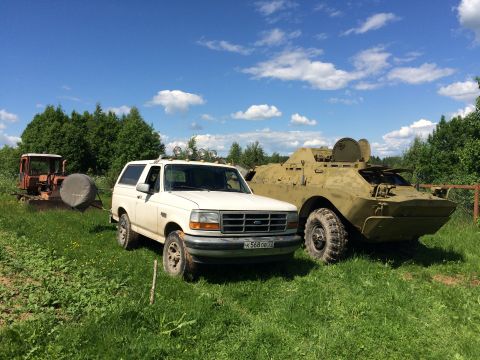 1993 Ford Bronco - A white 1993 Ford Bronco is parked in a grassy field next to a military armored vehicle under a blue sky with clouds.
