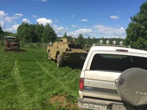 1993 Ford Bronco - A white SUV is parked in a grassy field next to a military armored vehicle, with a tractor visible in the distance.