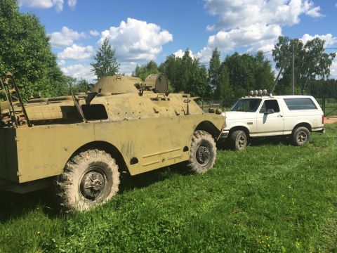 1993 Ford Bronco - A white 1993 Ford Bronco is parked on a grassy field next to a military armored vehicle under a cloudy sky.
