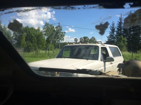 1993 Ford Bronco - A white 1993 Ford Bronco with auxiliary lights on its roof rack is parked outdoors, viewed through a dirty windshield.
