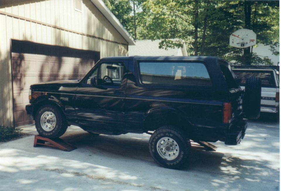Black Ford Bronco with rear wheel on a ramp parked beside a garage in a wooded yard